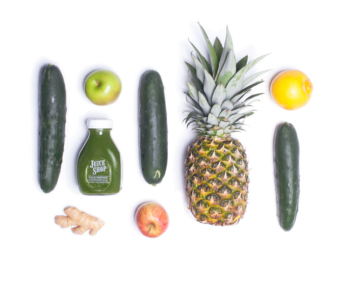 A flat lay arrangement of fresh ingredients used in Juice Shop’s cold-pressed green juice. The composition includes whole cucumbers, a ripe pineapple, green and red apples, fresh ginger, and a bright yellow lemon, symmetrically placed around a bottle of Juice Shop’s cold-pressed juice. The clean white background emphasizes the vibrant colors and natural freshness of the ingredients.