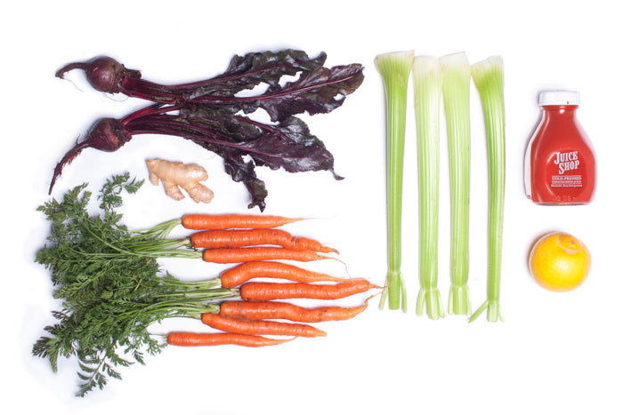A flat lay composition of fresh organic ingredients used in Juice Shop’s cold-pressed juice. The arrangement includes deep red beets with greens, vibrant orange carrots with tops, fresh ginger, crisp celery stalks, and a bright yellow lemon. A bottle of Juice Shop’s cold-pressed juice sits beside the ingredients, showcasing the final product.