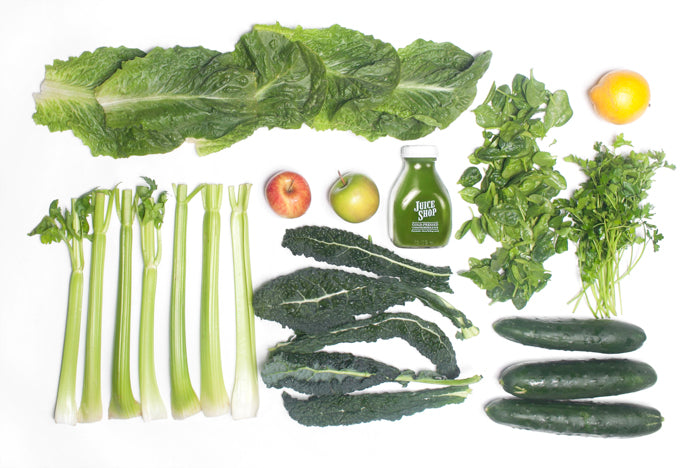 A flat lay display of fresh, organic ingredients used in Juice Shop’s cold-pressed green juice. The arrangement includes crisp romaine lettuce, leafy kale, fresh spinach, vibrant celery stalks, cucumbers, apples, a lemon, and a bunch of cilantro. A bottle of Juice Shop’s green juice is placed in the center, showcasing the final product made from these nutrient-rich ingredients.