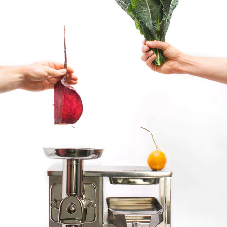 Hands holding beet and kale above a stainless steel juicer.
