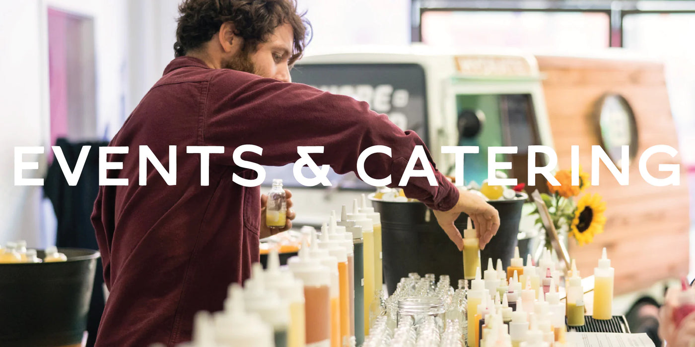 A Juice Shop team member prepares fresh juice samples at an event, carefully pouring vibrant juices into small bottles. The table is lined with an assortment of colorful juice-filled squeeze bottles and empty glasses, ready for serving. A Juice Shop-branded vehicle and sunflowers are visible in the background, enhancing the catering setup. Large white text overlay reads 'EVENTS & CATERING.