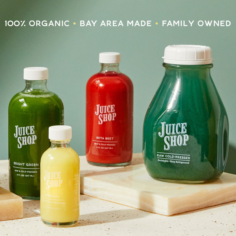 A collection of Juice Shop's organic, cold-pressed juices in glass bottles of various sizes, displayed on marble slabs against a green background. The juices come in vibrant colors, including green, red, yellow, and deep blue, with labels indicating flavors like Bright Green, Beta Beet, and Elderberry. Above the bottles, text reads: '100% Organic • Bay Area Made • Family Owned.