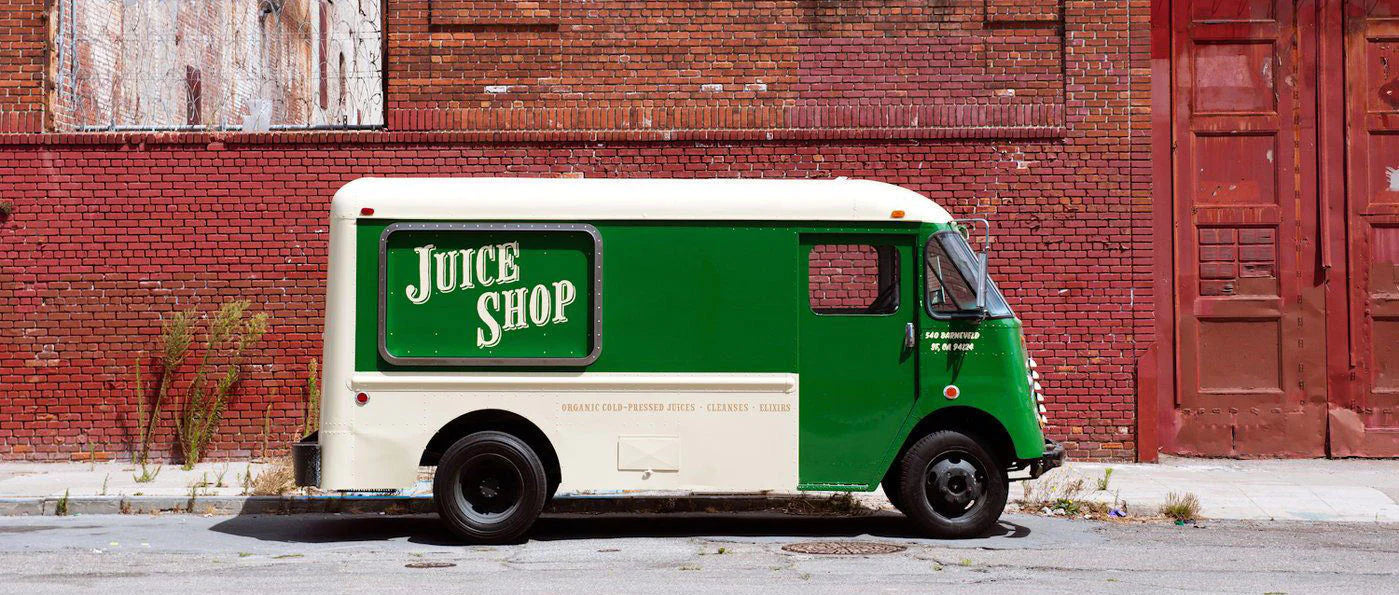 A vintage Juice Shop delivery truck parked on an urban street against a red brick wall with aged textures and overgrown weeds. The truck is painted in a two-tone green and cream design, featuring a large Juice Shop logo on the side panel. The vehicle has a retro aesthetic with rounded edges, a boxy frame, and black tires. The text 'Organic Cold-Pressed Juices • Cleanses • Elixirs' is subtly printed along the lower side of the truck. The background gives an industrial feel, enhancing the nostalgic charm 